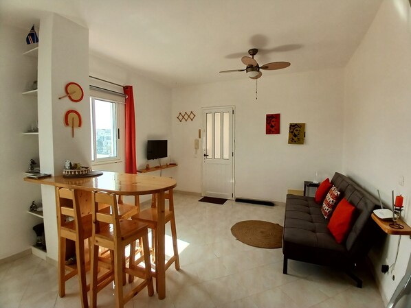 A living room with a sofa, a dining table with chairs, a ceiling fan, and a TV.