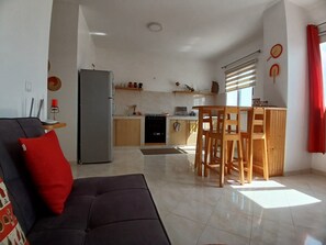 A modern living room with a sofa, a dining area with wooden chairs, a kitchen with a refrigerator, and a window with blinds.