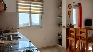A kitchen with a gas stove, sink, and window with blinds. There are wooden chairs and a TV on a stand. - The Yellow Flat - Sea View (Boa Vista)