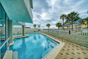 A swimming pool with a tiled deck, palm trees, and a building with a blue roof.