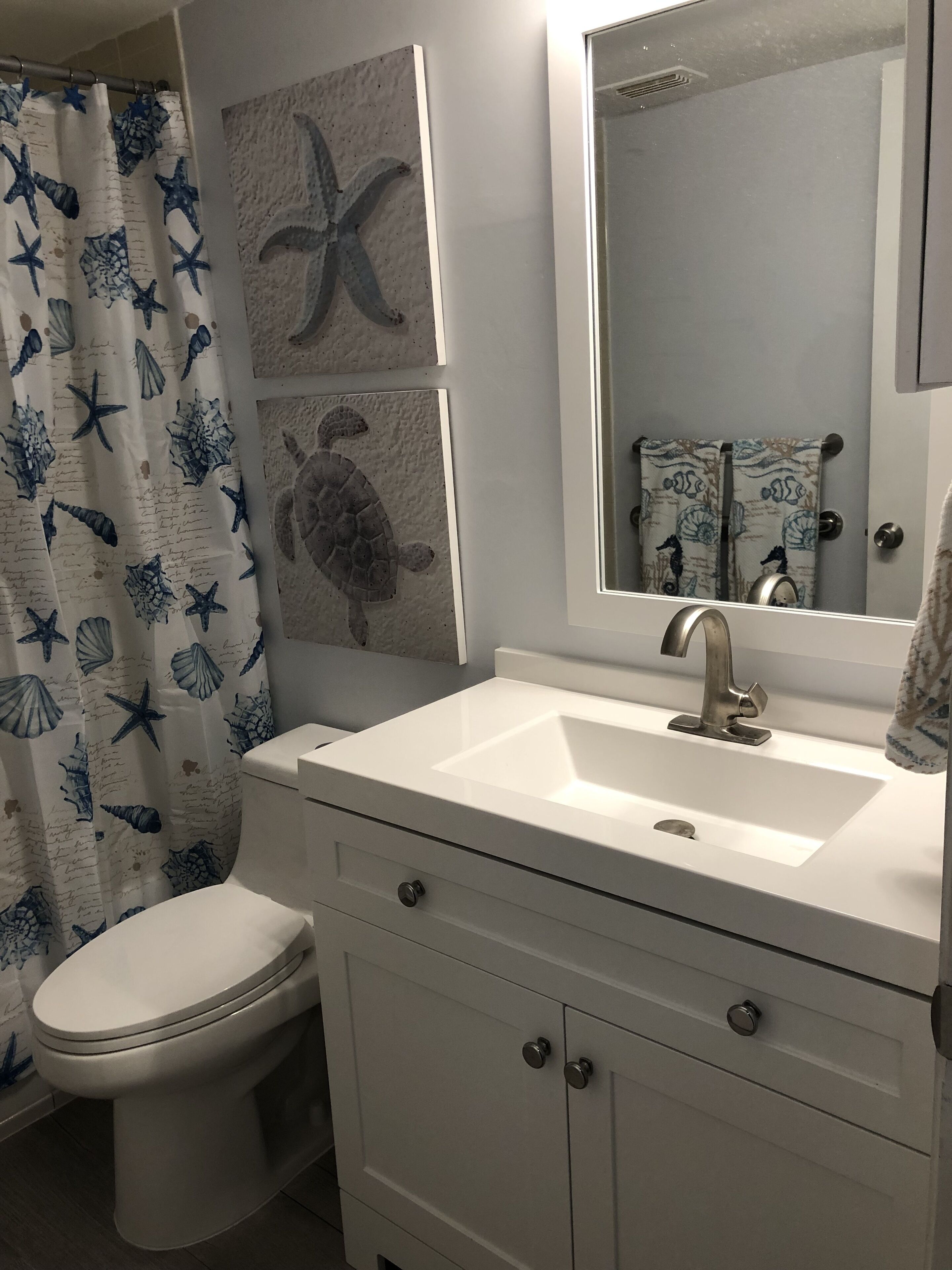 A bathroom with a white sink, a toilet, and a shower curtain with starfish and seashells. There are wall art pieces above the sink and a mirror above the sink.