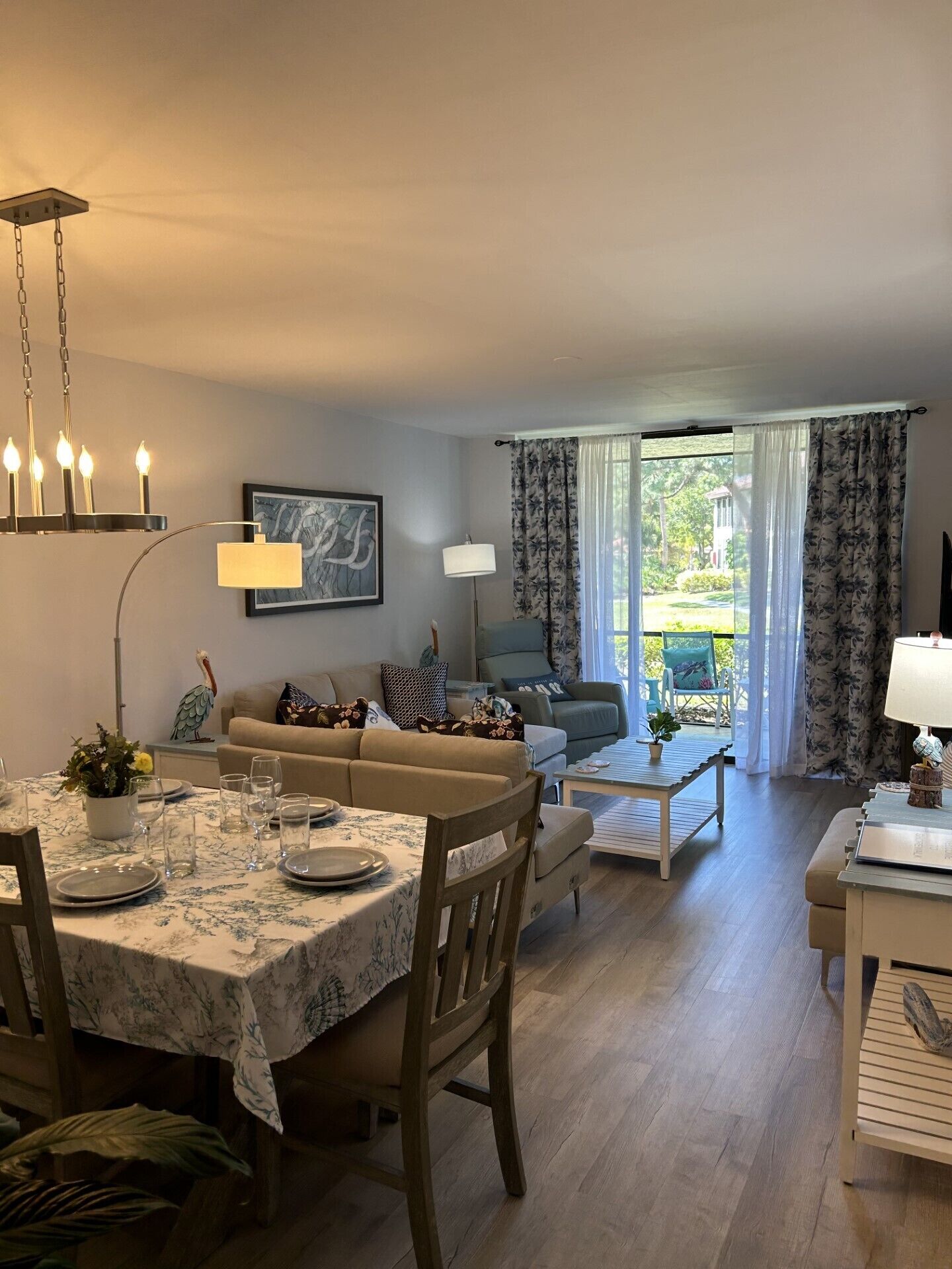 A dining area with a table set for four, a sofa, a coffee table, and a view of the outdoors through large windows.