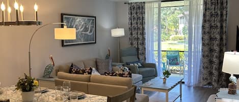 A dining area with a table set for four, a sofa, a coffee table, and a view of the outdoors through large windows.