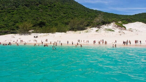 Ein Strand mit badenden und sonnenden Menschen, umgeben von einem bewaldeten Hügel.