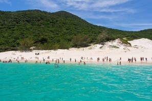 A beach with people swimming and sunbathing, surrounded by a forested hill. - Arraial do Cabo Casa Aluguel Economico (Arraial do Cabo)