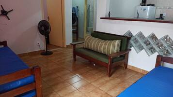 Una habitación con dos camas de madera, un sofá verde con un cojín a rayas, un ventilador de pie y una zona de cocina con un refrigerador y una cafetera.
