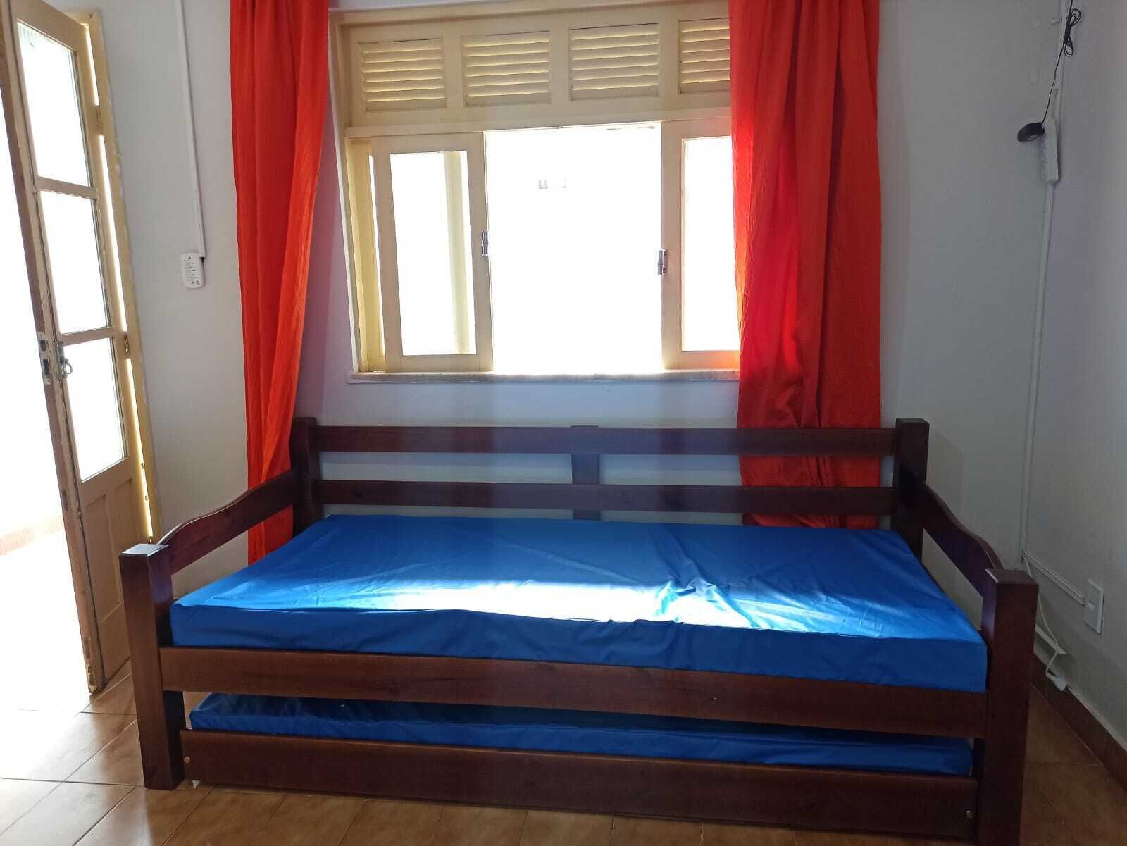 A wooden bed with a blue mattress in a room with a window and red curtains.