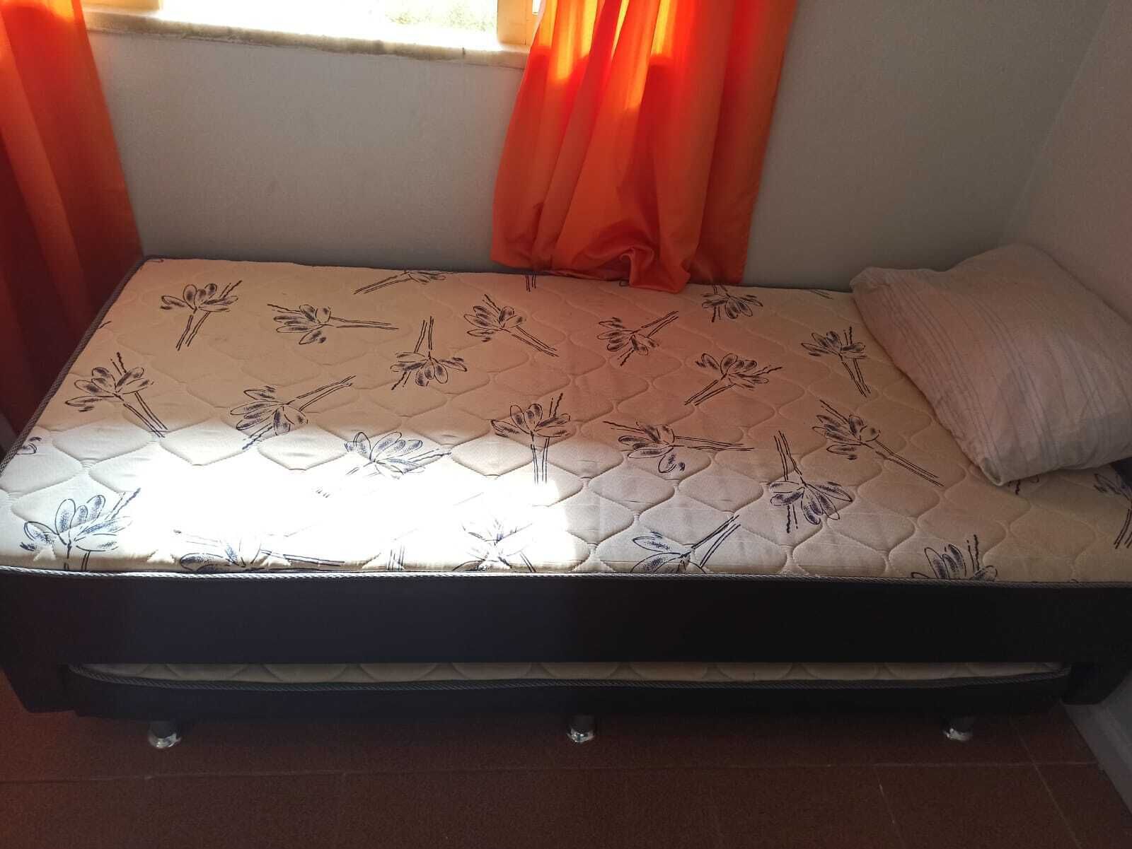 A single bed with a floral-patterned mattress in a room with a window and orange curtains.