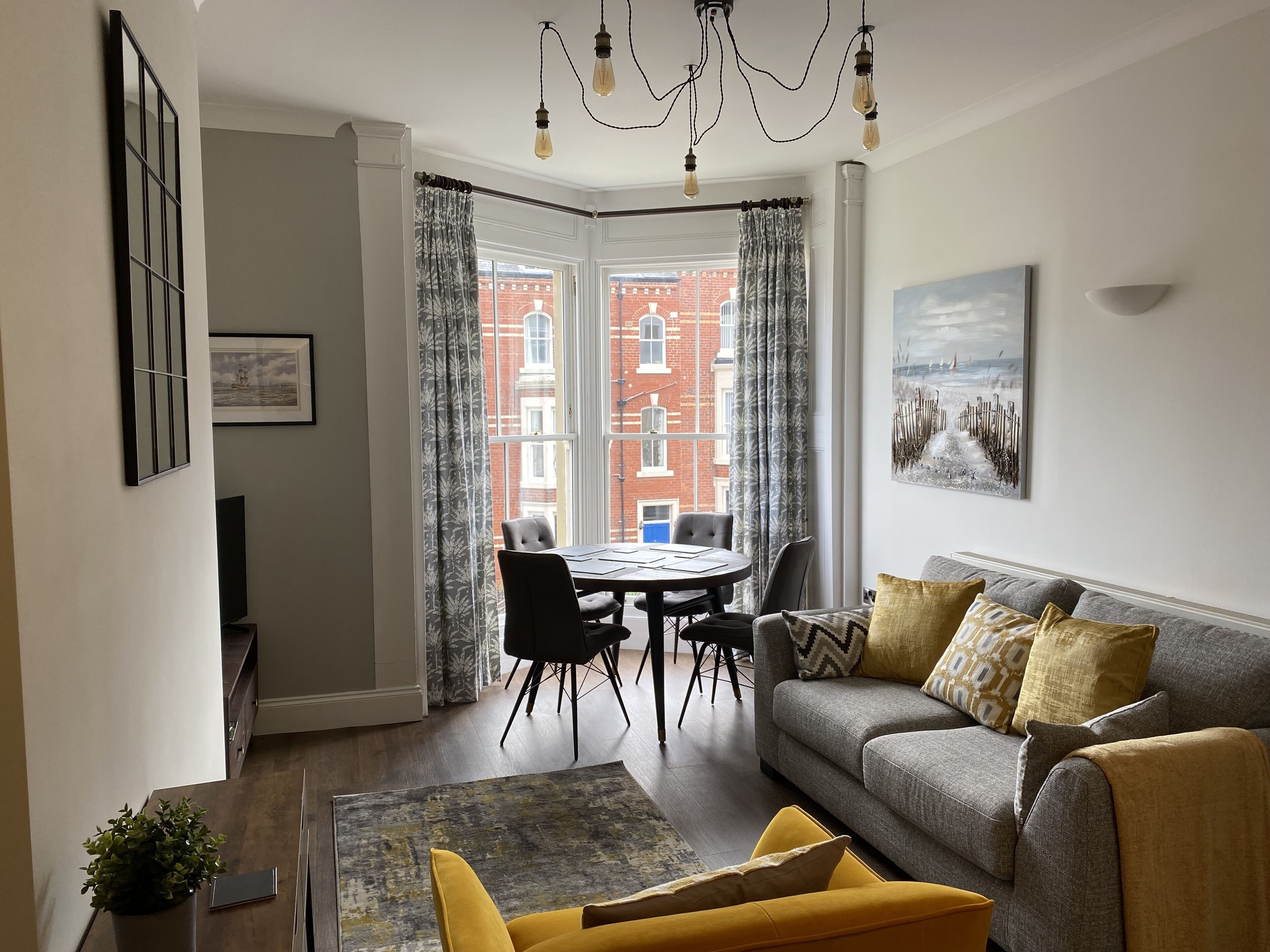 A modern living room with a grey sofa, yellow armchair, and a dining table with chairs. A large window offers a view of a red brick building.