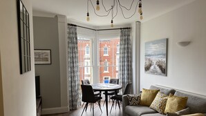 Een moderne woonkamer met een grijze bank, een gele fauteuil en een eettafel met stoelen. Een groot raam biedt uitzicht op een gebouw van rode baksteen.
