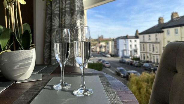Twee glaasjes water op een tafel bij een raam, met uitzicht op gebouwen en een weg.