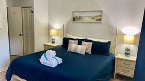 A neatly made bed with a dark blue bedspread, two bedside tables with lamps, a door, and a painting on the wall. - 162 The Greens Villa Walk to Pool, Beach (Hilton Head Island)
