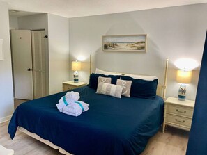 A neatly made bed with a dark blue bedspread, two bedside tables with lamps, a door, and a painting on the wall.