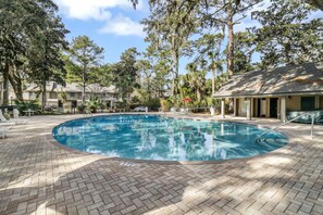 A swimming pool with a brick deck, surrounded by trees and a building with a covered area.