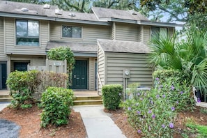 A two-story residential building with a front entrance, a small porch, and a landscaped garden with various plants and flowers.