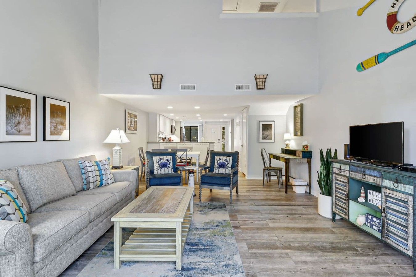 A modern living room with a sofa, chairs, a coffee table, and a TV. There is a kitchen in the background with cabinets and a dining area.