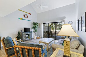 A spacious living room with a television, a coffee table, and a sofa. There is a plant and a lamp in the room.