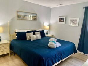 A neatly made bed with a blue bedspread and pillows, a bedside lamp, and a folded towel on the nightstand.