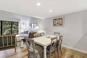 A dining area with a white table and chairs, a sofa with cushions, a clock, and framed artwork on the wall.