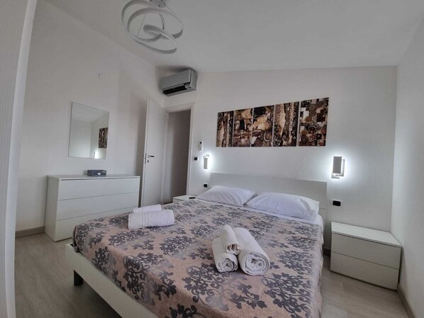 Une chambre à coucher comprenant un lit, une table de chevet, une commode, un meuble à tiroirs, un miroir, un appareil de climatisation et une œuvre d’art accrochée au mur.