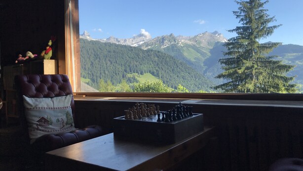 A room with a chessboard on a table, a window with a mountain view, and a plush chair with a decorative pillow.