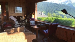 A cozy cabin interior with a large window offering a mountain view, a wooden ceiling, and a comfortable seating area with a table and chairs.