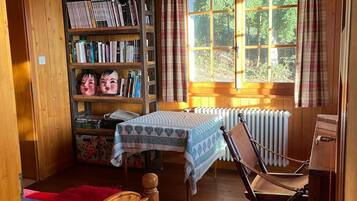 A cozy cabin room with a wooden floor, a bed, a desk, a chair, a bookshelf, and a window with curtains.