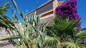 Un edificio con muro de piedra, balcón y varios plantas, incluyendo un arbusto grande con flores moradas.