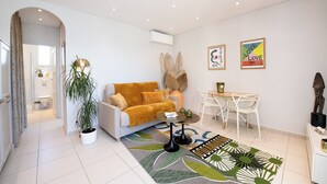 Una sala de estar moderna con un sofá, una mesa de centro y sillas. Hay una planta en maceta, obras de arte en la pared y un comedor con una mesa y sillas.