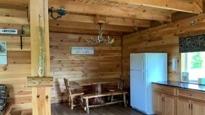 Interior rústico de una cabaña con una mesa de madera, un banco, un refrigerador blanco y un letrero que dice 'GATHER 'ROUND the table'.