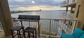 Un balcón con mesa y dos sillas, con vista a una masa de agua y un horizonte urbano distante.