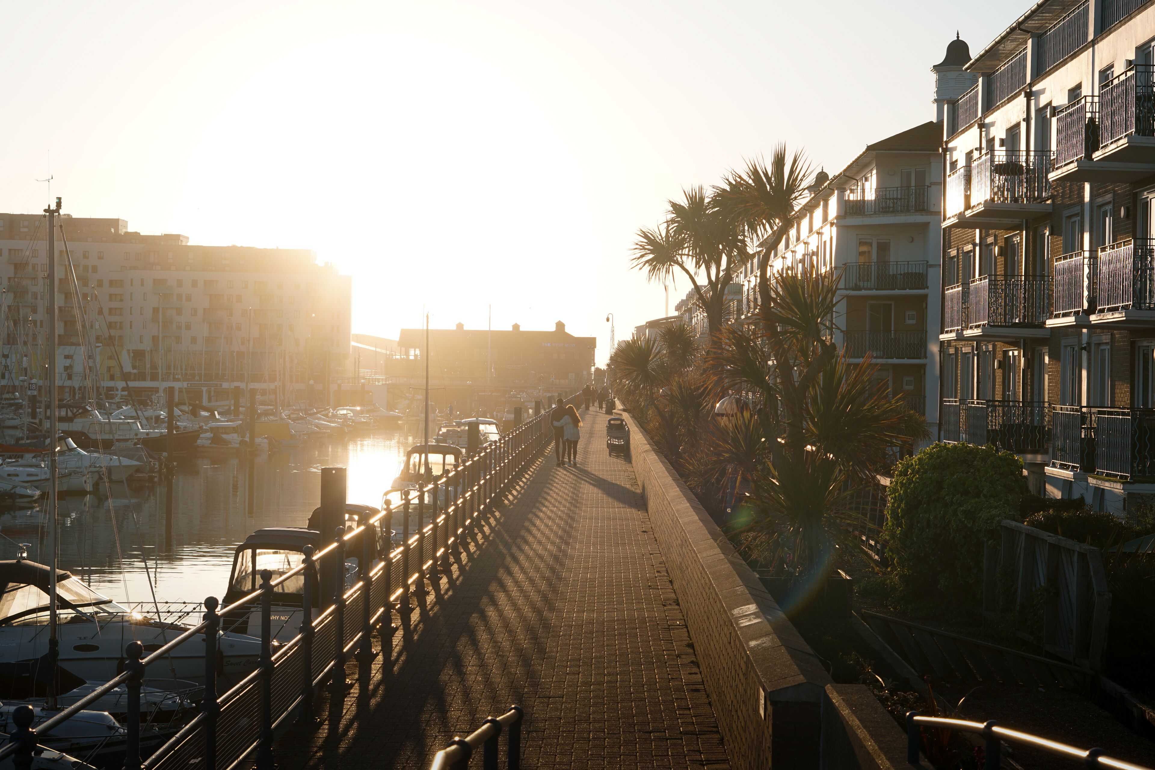A waterfront promenade with a brick walkway, boats, and buildings with balconies.