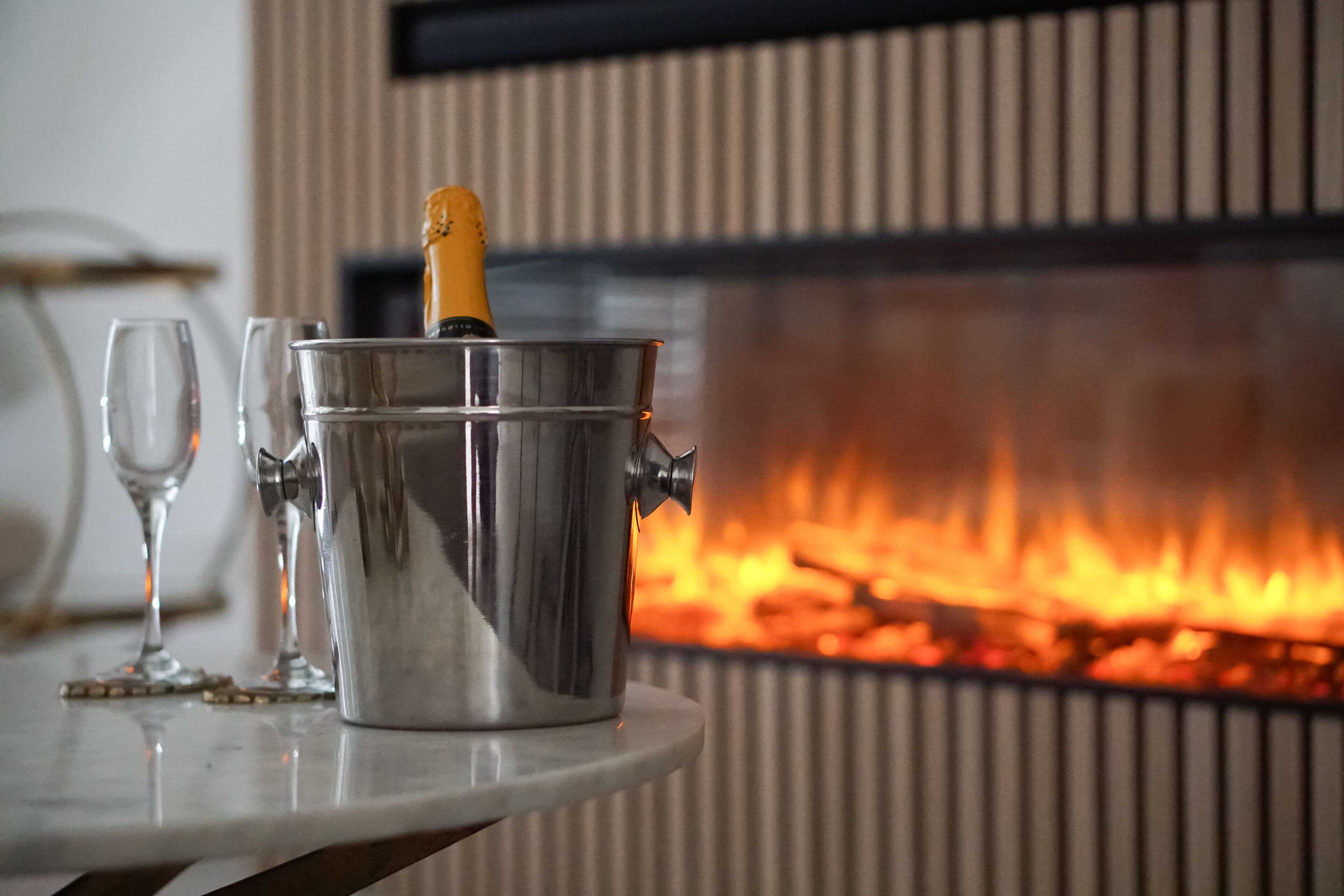 Una botella de champán en un cubo de metal sobre una mesa con dos copas de vino, frente a una chimenea con fuego.