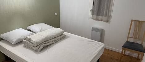 A single bed with white linens, a wooden chair, a radiator, and a window with a curtain.