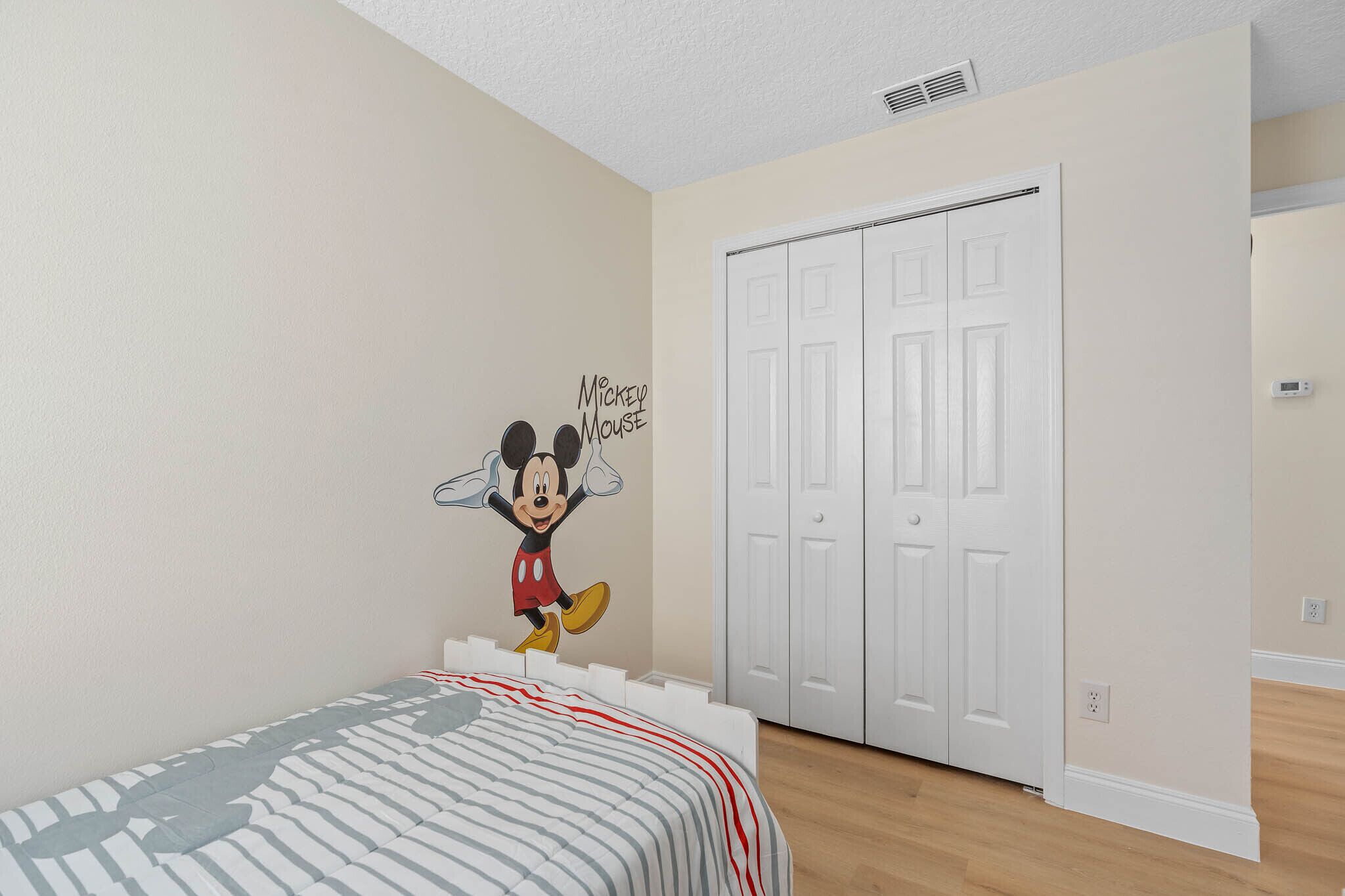A bedroom with a bed, a Mickey Mouse wall decal, and a white wardrobe.