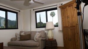 A room with a sofa, a lamp, a wooden cabinet, and large windows with a view of palm trees and mountains. - Lake Front (Ajijic)