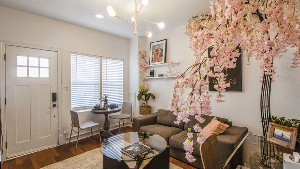 A living room with a sofa, a glass coffee table, a chair, a bookshelf, and a large window with blinds.