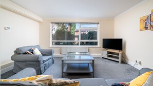 A living room with a sofa, a coffee table, a TV, and a window with a view of buildings and trees. - Spacious Pet Friendly 2-bed Apartment in Redhill (Redhill)