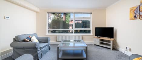 A living room with a sofa, a coffee table, a TV, and a window with a view of buildings and trees.