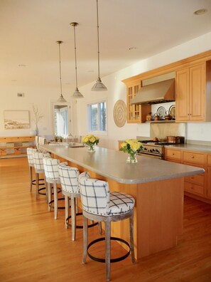 A modern kitchen with a central island, bar stools, pendant lighting, wooden cabinets, and a view of the outdoors.