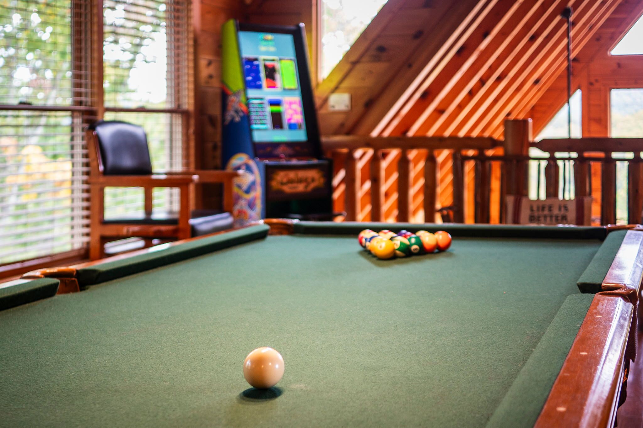 A pool table with balls, a pool cue, and a pool table in a room with wooden walls and a window.