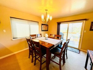 A dining room with a wooden table and chairs, a chandelier, and large windows with blinds.