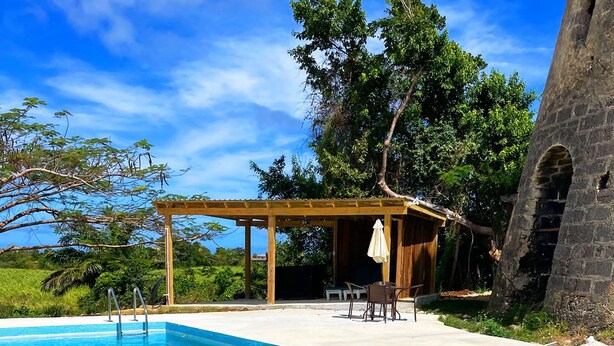A clear blue pool with a wooden pergola and chairs, surrounded by lush greenery and a stone structure. - Historic sea view 1 bed apartment in restored stone house (Four Hill)