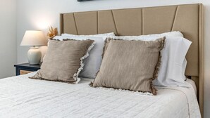 Una cama bien hecha con un cabecero color beige, sábanas blancas y dos almohadas beige con bordes deshilachados.
