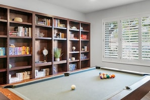 Une table de billard avec une boule blanche et des boules de couleurs, entourée d’étagères en bois remplies de livres et d’objets décoratifs, et une fenêtre avec des stores blancs.