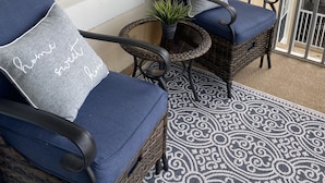 Un balcon avec deux fauteuils en osier, dont l’un est agrémenté d’un coussin et d’un oreiller orné de la mention « chez soi, quel bonheur », ainsi qu’une petite table sur laquelle est posée une plante.
