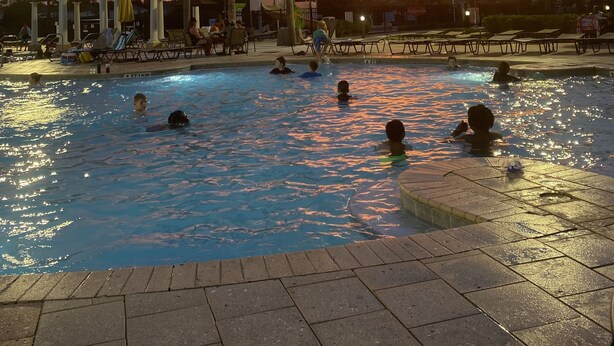 Une piscine où l’on voit des personnes dans l’eau, entourée de palmiers, avec un coucher de soleil en arrière-plan.