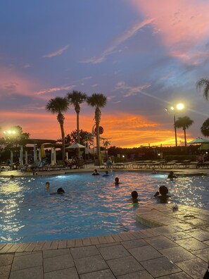 A swimming pool with people in the water, surrounded by palm trees and a sunset in the background. - Be our Guest - 3 bedroom, sleeps 7 + Condo w/ pool view by Disney and Universal (Kissimmee)