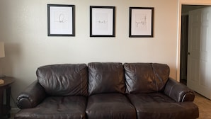 Un canapé en cuir marron se trouve dans une pièce dont un mur est recouvert d’un revêtement texturé blanc. Trois panneaux encadrés sont accrochés au mur, et portent les mots « soyez », « nos » et « invités ».