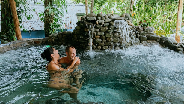 A couple in a clear indoor pool with a waterfall feature and a stone wall. - Oasis Diverse Adult Retreat. (Quepos)